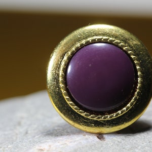 May include: A gold-coloured button with a purple centre. The button has a decorative, rope-like border around the edge.