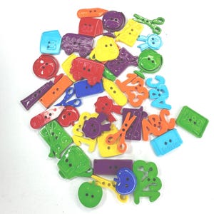 May include: A collection of assorted plastic buttons in various bright colours and school-themed shapes, including scissors, rulers, apples, globes, buses, and letters. These decorative fasteners feature two or four holes for sewing onto garments.