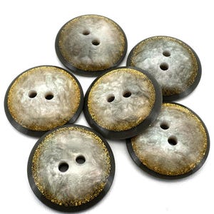 Op de afbeelding: Zes ronde knopen met een parelmoer centrum en een donkergrijze rand. De knopen hebben twee gaatjes en een gouden glitterrand. De knopen zijn gerangschikt op een witte achtergrond.