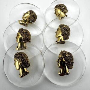 May include: A collection of clear, circular buttons with a gold-colored profile of a woman's face. The buttons are made of clear plastic and have a vintage aesthetic. The gold-colored face is detailed with hair and facial features.