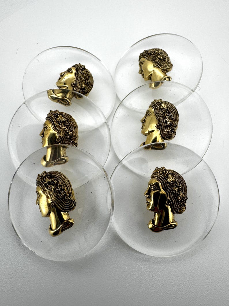 May include: A collection of clear, circular buttons with a gold-colored profile of a woman's face. The buttons are made of clear plastic and have a vintage aesthetic. The gold-colored face is detailed with hair and facial features.