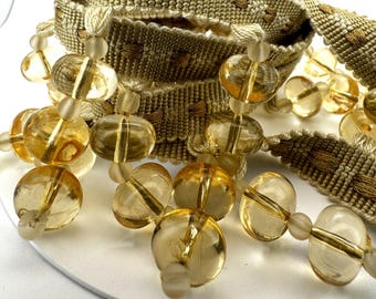 以下が含まれることがあります： ベージュとゴールドの織り込まれたトリムのクローズアップ。フリンジとクリアアンバーガラスビーズのクラスターがあります。