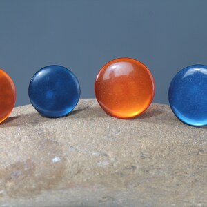 Op de afbeelding: Vier ronde, doorschijnende cabochons in tinten oranje en blauw zijn gerangschikt op een stenen oppervlak. De cabochons hebben een glanzende afwerking en zijn gelijkmatig verdeeld, met twee oranje en twee blauwe.