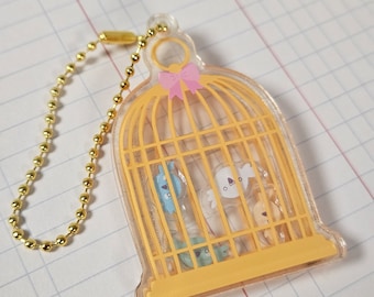 May include: A yellow birdcage keychain with a pink bow and a gold chain. The cage is made of clear plastic and has small, colorful birds inside.