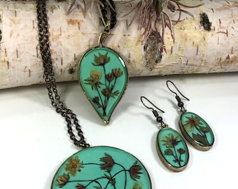 May include: A set of jewellery featuring a teal pendant and earrings with pressed flowers. The pendant is a round shape with a bronze frame and the earrings are oval shaped with bronze frames.