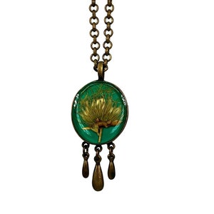Puede incluir: Un collar con colgante de estilo vintage con una flor dorada encapsulada en resina ovalada verde. El colgante cuelga de una cadena de color bronce y tiene tres colgantes en forma de lágrima de color bronce. El collar está sobre un fondo blanco.