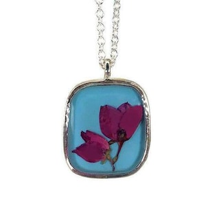 Puede incluir: Collar plateado con colgante cuadrado. El colgante tiene un fondo azul claro con dos flores de color rosa oscuro. El colgante está enmarcado en plata y cuelga de una delicada cadena. Un accesorio de temática floral.