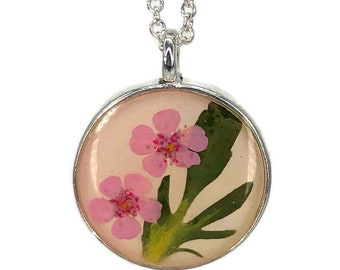 Op de afbeelding: Een zilveren ketting met een ronde hanger met twee geperste roze bloemen en groene bladeren ingekapseld in helder hars.