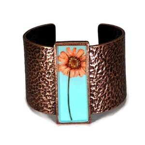 May include: Wide copper cuff bracelet with a hammered texture. A rectangular turquoise resin inlay features a pressed orange flower. The cuff has a black interior.