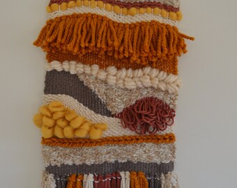Puede incluir: Un tapiz de pared tejido con una variedad de texturas y colores, incluyendo marrón, beige, naranja y rojo. El tapiz tiene un borde de flecos y está hecho con hilo.