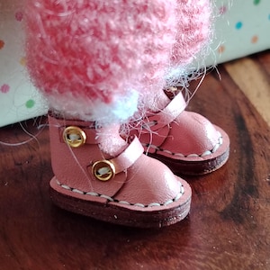 Könnte beinhalten: Nahaufnahme von Miniatur-Lederstiefeln in Rosa mit goldfarbenen Schnallen und weißen Nähten. Die Stiefel sind mit einem flauschigen rosa Kleidungsstück mit weißem Besatz kombiniert. Die Stiefel stehen auf einer Holzoberfläche.