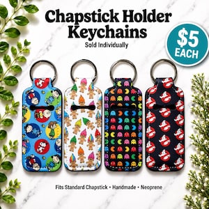May include: Four chapstick holder keychains with different patterns: a blue one with cartoon characters, a white one with trolls, a black one with Pac-Man, and a black one with Ghostbusters logos. The text "Chapstick Holder Keychains" is at the top.