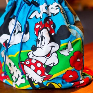 May include: A blue drawstring bag featuring Minnie Mouse and Mickey Mouse. The bag has a blue drawstring closure and a colorful design with the word "MICKEY" in white. The bag is made of a soft, flexible material.