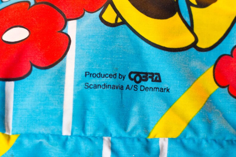 May include: Close-up of a blue fabric with a floral design in red and yellow. The text "Produced by COBRA Scandinavia A/S Denmark" is printed in black.