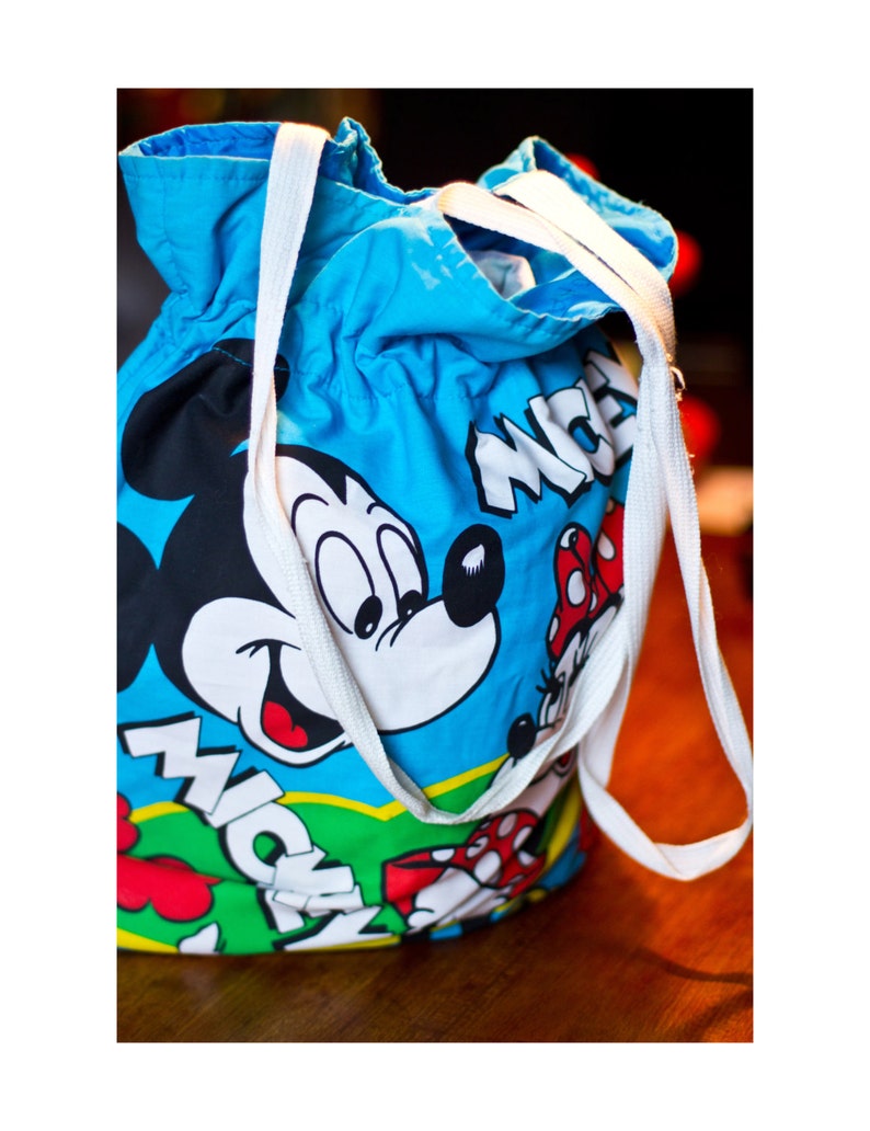 May include: A blue drawstring bag featuring Mickey Mouse and Minnie Mouse graphics. The bag has white straps and the word "Mickey" in white block letters. The bag is made of a lightweight fabric and is suitable for carrying items.