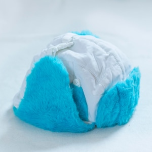 Puede incluir: Sombrero de invierno blanco con orejeras de piel sintética azul. El sombrero está hecho de un tejido acolchado y tiene un diseño sencillo.
