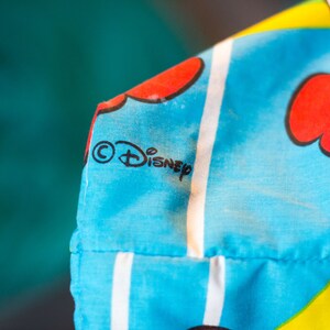 May include: Close-up of a section of a blue fabric with a floral pattern and white stripes. The fabric features the Disney logo in black. Red and white flowers are also visible.