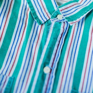 Puede incluir: Una camisa de botones a rayas verdes, blancas, azules y rojas con una etiqueta de Calvin Klein. La camisa tiene un cuello abotonado y un bolsillo en el lado izquierdo.