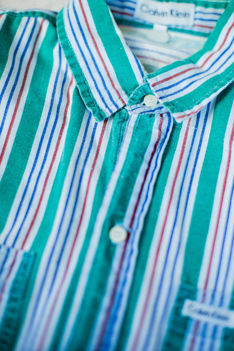 Puede incluir: Una camisa de botones a rayas verdes, blancas, azules y rojas con una etiqueta de Calvin Klein. La camisa tiene un cuello abotonado y un bolsillo en el lado izquierdo.