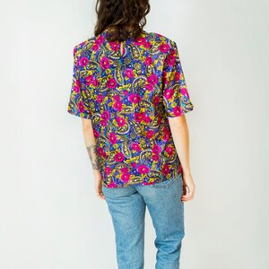 K&ouml;nnte beinhalten: Eine kurz&auml;rmelige Bluse mit floralem Druck in Lila, Rosa, Gelb und Blau mit einem runden Ausschnitt.