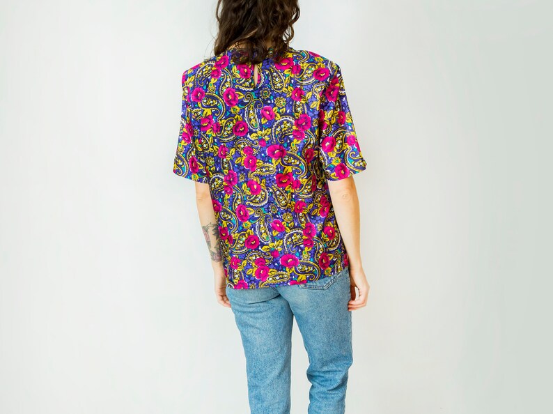 K&ouml;nnte beinhalten: Eine kurz&auml;rmelige Bluse mit floralem Druck in Lila, Rosa, Gelb und Blau mit einem runden Ausschnitt.