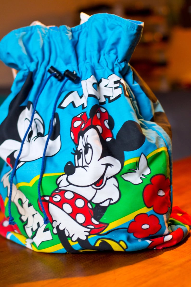 May include: A blue drawstring bag featuring Minnie Mouse and Mickey Mouse. The bag has a blue drawstring closure and a colorful design with the word "MICKEY" in white. The bag is made of a soft, flexible material.