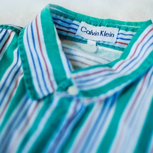 Puede incluir: Una camisa de botones a rayas verdes y blancas con una etiqueta de Calvin Klein. La camisa tiene cuello y cierre de botones.