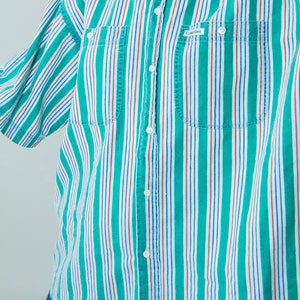 Puede incluir: Una camisa de botones a rayas con rayas verdes, blancas, rojas y azules. La camisa tiene cuello, cierre de botones y dos bolsillos en el pecho. La camisa tiene el texto "Calvin Klein" en el bolsillo izquierdo del pecho.