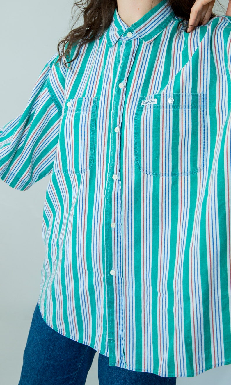 Puede incluir: Una camisa de botones a rayas con rayas verdes, blancas, rojas y azules. La camisa tiene cuello, cierre de botones y dos bolsillos en el pecho. La camisa tiene el texto "Calvin Klein" en el bolsillo izquierdo del pecho.