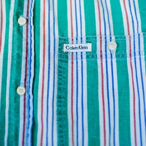 Puede incluir: Una camisa de botones a rayas con un patr&oacute;n verde, blanco, rojo y azul. La camisa tiene un bolsillo en el pecho izquierdo con una etiqueta "Calvin Klein".
