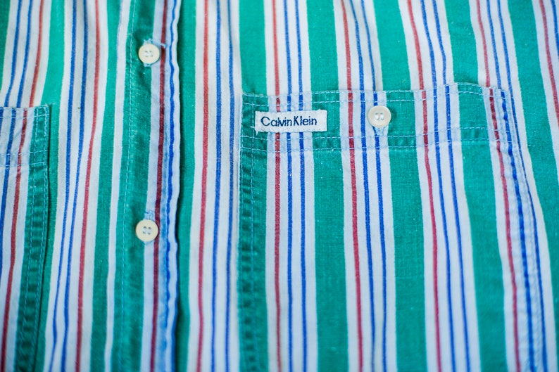Puede incluir: Una camisa de botones a rayas con un patr&oacute;n verde, blanco, rojo y azul. La camisa tiene un bolsillo en el pecho izquierdo con una etiqueta "Calvin Klein".
