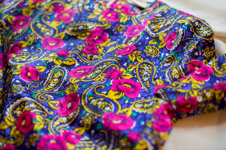 K&ouml;nnte beinhalten: Nahaufnahme eines blauen Stoffes mit Paisley-Muster. Der Stoff ist mit rosa Blumen und gelben Akzenten verziert.