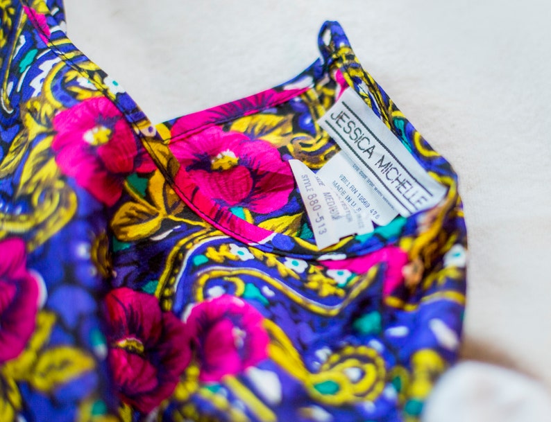 K&ouml;nnte beinhalten: Eine blaue, rosa, gelbe und gr&uuml;ne Seidenbluse mit floralem Muster und einem wei&szlig;en Etikett mit der Aufschrift "Jessica Michelle" und "Style 680-513".