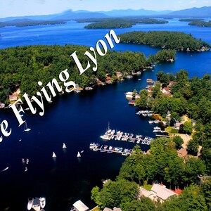 Puede incluir: Vista aérea de un lago con varias islas y un pequeño pueblo en la costa. El agua es de un azul profundo y el cielo está despejado. El texto "The Flying Lyon" está superpuesto sobre la imagen.