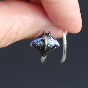 May include: A silver ring with a small, detailed resin stingray charm. The stingray is blue, black, and gold with a glittery finish.