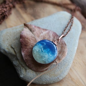 May include: A round pendant necklace with a copper chain. The pendant features a blue and white resin design, resembling ocean waves. The necklace is displayed on a gray stone with a brown leaf.