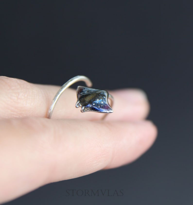 May include: A silver ring with a black and blue stingray design. The ring is on a finger.