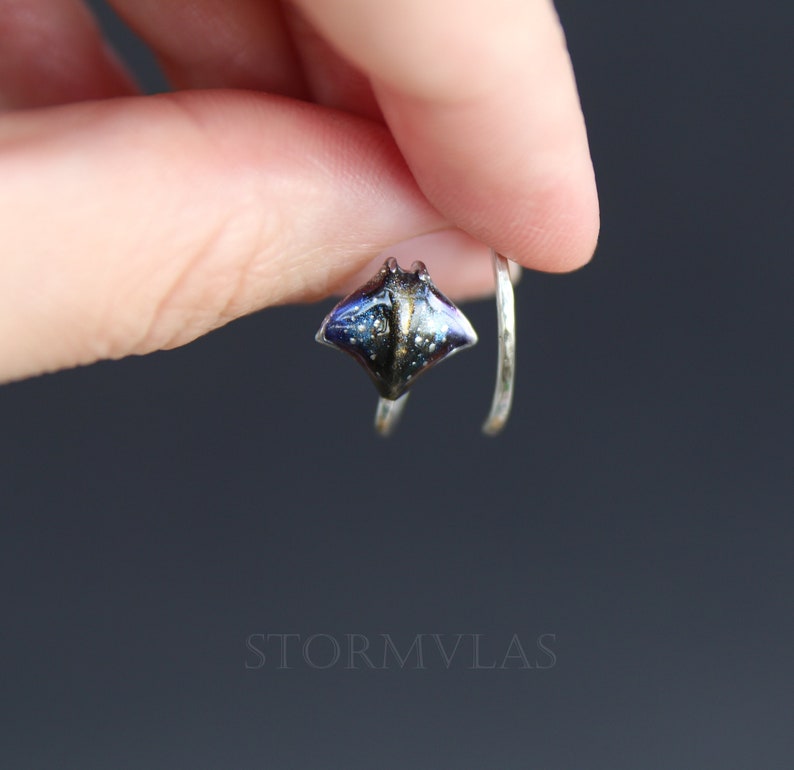 May include: A silver ring with a small, detailed resin stingray charm. The stingray is blue, black, and gold with a glittery finish.