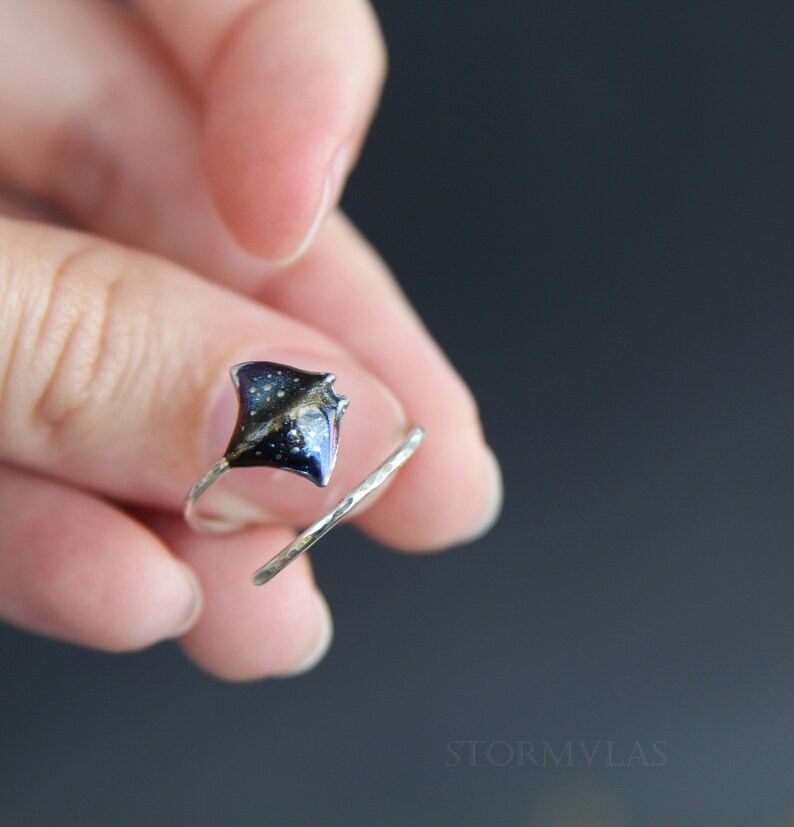 May include: A silver ring with a black and blue stingray charm. The stingray has a galaxy-like design with white and gold glitter.