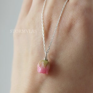 May include: A delicate silver chain necklace with a pink and green flower pendant. The flower is a realistic depiction of a closed bud.