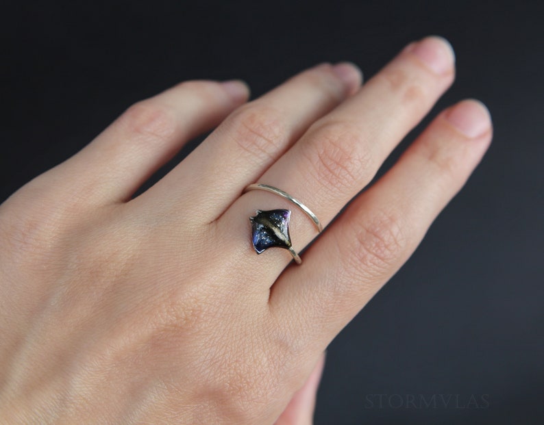 May include: A silver ring with a blue and black stingray design. The ring is worn on a person's finger.