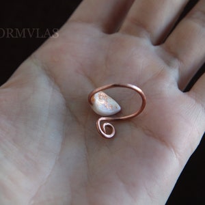K&ouml;nnte beinhalten: Ein Ring aus Kupferdraht mit einem wei&szlig;en und goldenen Opal-Stein. Der Ring befindet sich auf einer Hand.