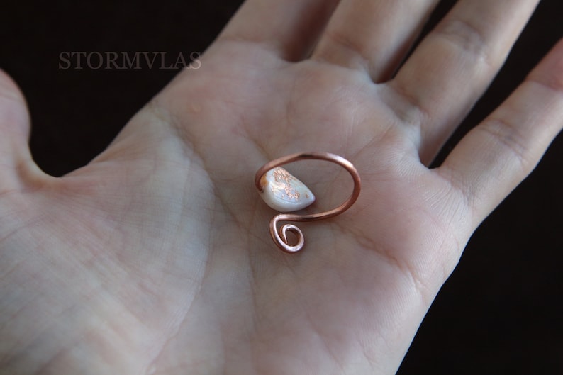 K&ouml;nnte beinhalten: Ein Ring aus Kupferdraht mit einem wei&szlig;en und goldenen Opal-Stein. Der Ring befindet sich auf einer Hand.