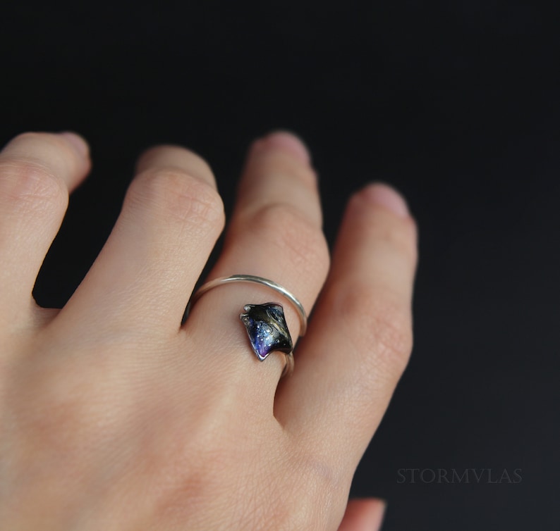 May include: A silver ring with a dark blue and black resin inlay shaped like a manta ray. The ring is worn on a person's finger.