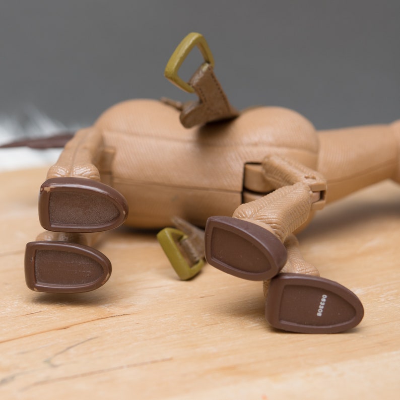 May include: A brown plastic toy horse with a saddle and stirrups. The horse is lying on its side with its legs in the air. The horse's hooves are brown and have the word "RODEO" printed on them.