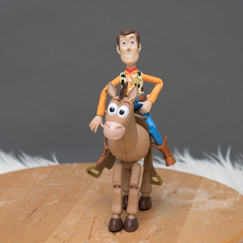 May include: A plastic toy figure of Woody from Toy Story riding a brown plastic horse. Woody is wearing his signature cowboy hat, vest, and jeans. The horse has a saddle and stirrups.