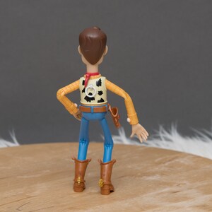 May include: A plastic action figure of Woody from the Toy Story franchise. He is wearing his signature cowboy hat, vest, and jeans. He is standing with his arms outstretched and his legs slightly bent.