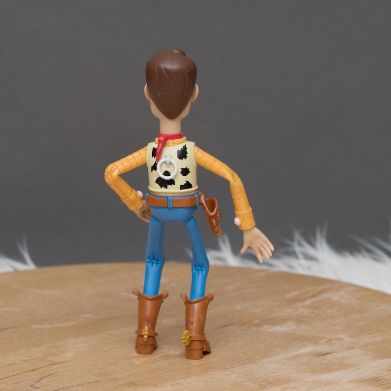 May include: A plastic action figure of Woody from the Toy Story franchise. He is wearing his signature cowboy hat, vest, and jeans. He is standing with his arms outstretched and his legs slightly bent.