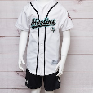 May include: A white baseball jersey with black trim and the word "Marlins" in black and teal lettering. The jersey features a small teal, black, and white logo of a marlin fish on the left chest. The jersey is paired with black shorts with a white "A" logo on the left leg.