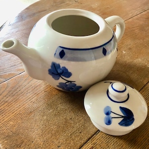 Puede incluir: Tetera de cerámica blanca con un diseño floral azul y tapa a juego. La tetera tiene un pico, un asa y una banda azul con forma de diamante. La tapa tiene un pomo y un patrón floral azul similar.